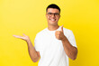 © luismolinero - Young handsome man over isolated yellow background holding copyspace imaginary on the palm to insert an ad and with thumbs up