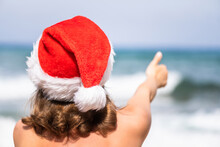 Christmas Sea Holiday. Back View Of Happy Woman In Santa Hat Showing Thumb Up And Relaxing On Paradise Beach Island Getaway. New Year