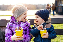 Two Cute Adorable Siblings Children Sitting On Bench Drink Delicious Yummy Hot Chocolate, Tea Cocoa From Paper Cups During Walk At City Street Park Or Backyard Outdoors. Brother And Sister Enjoy Fun