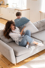 Wall Mural - Overjoyed single mom and little son have fun together play childish pillow fight in living room. Cheerful happy mother engaged in funny family activity with preschool boy on vacation or home education