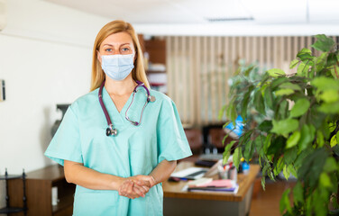 Wall Mural - Portrait of friendly female doctor in mask wearing uniform standing with stethoscope in modern clinic