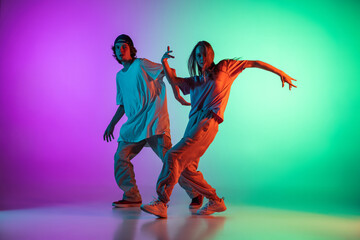 Wall Mural - Young hip-hop dancers, stylish emotive girl and boy in action and motion in casual sports youth clothes on gradient multi colored background at dance hall in neon light.