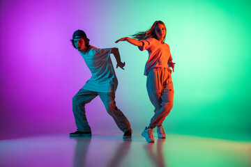 Wall Mural - Stylish young hip-hop dancers, emotive girl and boy in action and motion in casual sports youth clothes on gradient multi colored background at dance hall in neon light.