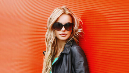 Stylish portrait of beautiful blonde woman wearing a black rock jacket and sunglasses on red background