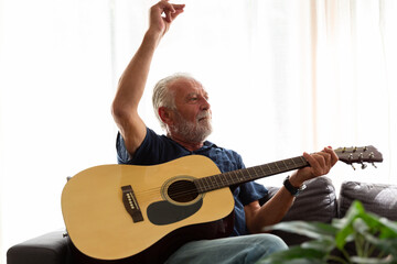 Cheerful senior man playing guitar while sitting on sofa at home. Elderly man learning guitar on holiday. people, quarantine, holiday concept