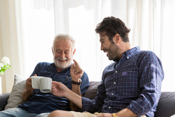 Wall Mural - Senior father with adult son talking and drinking coffee while sitting on sofa at home with happy.