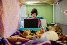 A Boy Sits In Bed With Knit Hat Doing Schoolwork On Computer With Cat