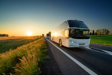 White double-decker bus traveling on the asphalt road in rural landscape at sunset