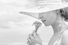 Portrait Of Young Beautiful Cheerful Woman Wearing Straw Sun Hat, Smelling Small Bouquet Of Yellow Wild Florets, Against Blue Summer Sky
