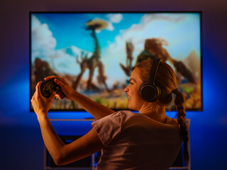 Wall Mural - Gamer girl sits in a room in front of a large computer monitor. She has headphones on her head and a joystick in her hands. Close-up. Video games, day off, hobby, quarantine.