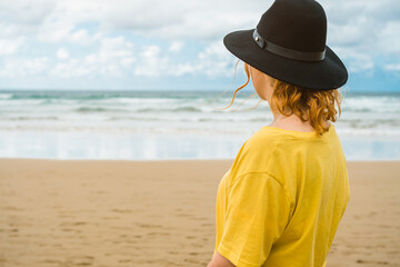 closeup of unrecognizable woman in yellow shirt and black hat standing on the beach looking at the sea