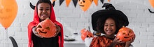 cheerful african american kids in halloween costumes holding carved pumpkins at home, banner