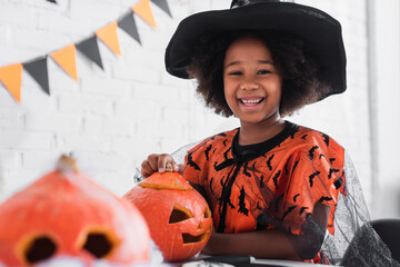 happy african american girl in witch halloween costume near caved pumpkins