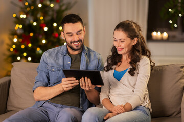 Sticker - technology, holidays and people concept - happy couple using tablet computer at home in evening over christmas tree lights on background