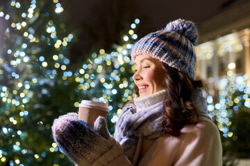 Sticker - winter holidays, hot drinks and people concept - happy smiling young woman drinking takeaway coffee over christmas tree lights outdoors