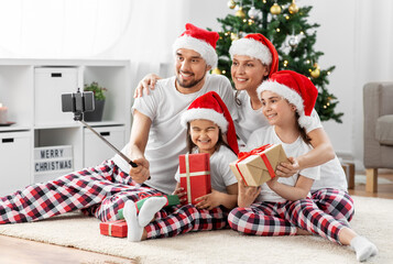 Canvas Print - family, winter holidays and people concept - happy mother, father and two daughters in santa hats sitting under christmas tree at home and taking selfie with smartphone