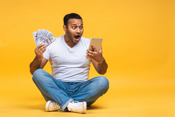 Portrait of african american indian black young man sitting on the floor holding dollar banknotes isolated over yellow background. Using tablet computer.