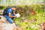 Fototapeta  - Biologist or botanist recording information about small tropical plants in forest. The concept of hiking to study and research botanical gardens by searching for information.