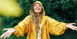 Positive young blonde woman smiling wearing yellow raincoat during the rain in the park. Cheerful female enjoying the rain outdoors. A beautiful woman catching the raindrops with arms wide open.