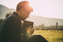 Man Praying On The Holy Bible In A Field During Beautiful Sunset.male Sitting With Closed Eyes With The Bible In His Hands, Concept For Faith, Spirituality, And Religion.