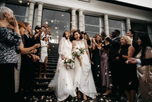 Candid Shot Of Two Female Lesbian LGBT Brides Walking Down The Stairs During Their Wedding Ceremony As Guests Throwing Rose Petal