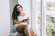 © F8  \ Suport Ukraine - Young woman sitting on the windowsill and writing message on smartphone at home