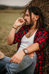 Poster - Young handsome Serbian male in a flannel shirt and ripped jeans sitting leaned on a haystack