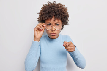 Poster - Serious African American woman with curly hair looks attentively at camera points directly at camera wears big transparent glasses blue turtleneck isolated over white background. Hey you listen me