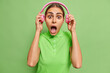 © WHstudio Leushin N - Scared young European woman puts off stereo headphones stares bugged eyes wide opened mouth cannot believe in shocking news dressed casually isolated over vivid green background. Monochrome shot