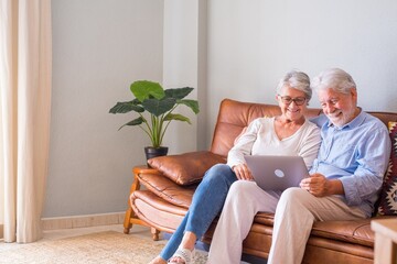 Cheerful senior couple using laptop while sitting on sofa and smiling. Elderly happy couple relaxing while surfing on laptop sitting in living room. Old couple watching media content using laptop