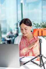 Smiling young dark-haired businesswoman with glasses sitting in front of a laptop screen in a cafe on a summer day and having video conference call via smartphone.Business,e-learning,freelance  concep