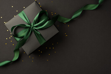 Wall Mural - Top view photo of stylish black giftbox with green satin ribbon bow and golden confetti on isolated black background with empty space