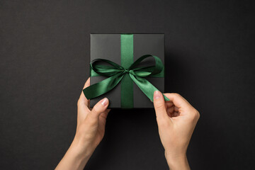 First person top view photo of female hands unpacking black giftbox with green satin ribbon bow on isolated black background