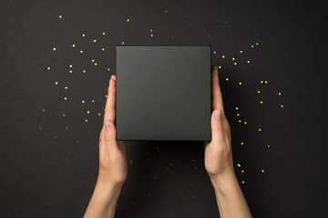 Wall Mural - First person top view photo of woman hands holding black giftbox and golden sequins on isolated black background with copyspace