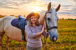Portrait of young cute happy joyful satisfied smiling woman hugging and stroking beautiful palomino blond horse at meadow at sunset