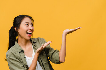 Portrait of young Asian lady smiling with cheerful expression, shows something amazing at blank space in casual clothing and standing isolated over yellow background. Facial expression concept.