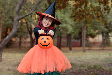 Wall Mural - Halloween trick or treat!Happy girl wearing a witch's hat, with a pumpkin handbag on the background of an autumn park. Funny kid in carnival costumes outdoors.Happy Halloween!