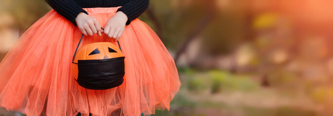 Wall Mural - Halloween trick or treat! A girl in a witch costume holds a bucket for sweets with a medical mask on the background of an autumn park. Halloween with safety measures from Covid-19. Banner