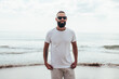 © San4ezz007 - Young bearded hipster guy with beard in white t-shirt and sunglasses at the tropical beach. Mock-up for print. T-shirt template.