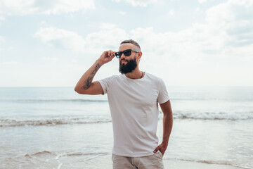 Wall Mural - Young bearded hipster guy with beard in white t-shirt and sunglasses at the tropical beach. Mock-up for print. T-shirt template.