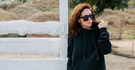 Beautiful young adult woman in black hoodie or sweatshirt and sunglasses against the background of a wooden fence and nature. Mock-up for print. Space for your logo or design.