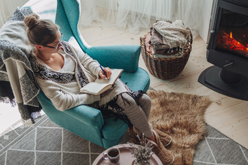 Young woman write in notebook sitting in armchair by fireplace