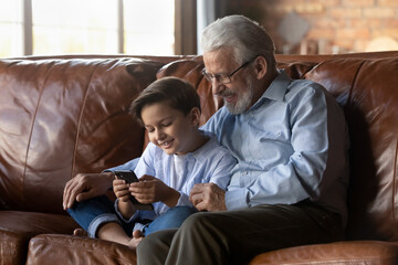 Wall Mural - Smiling grandfather with little grandson using phone, sitting on couch, happy grandpa in glasses with 8s boy kid watching funny video or cartoon, playing mobile device game, looking at screen