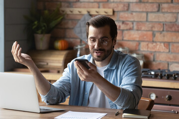 Wall Mural - man in glasses holding smartphone, chatting online by video call, sitting at table with laptop, smil