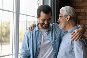 Wall Mural - Close up happy mature father and grownup son hugging cuddling enjoying tender moment, standing at home, smiling young man with senior dad in glasses having fun, two generations good relationship