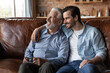 Happy dreamy two generations of men looking in distance, hugging, sitting on couch at home together, smiling senior dad with grownup son visualizing, dreaming of good future, enjoying weekend