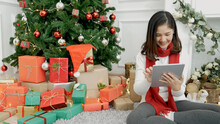 Asian Woman Using Smart Tablet Sit At Christmas Tree Xmas Present Gift Box In Cozy Living Room. Woman Hand Touch Screen Smile Shopping Online, Happiness Time Christmas Holiday Winter Season Festive