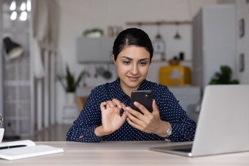 Wall Mural - Confident Indian businesswoman using smartphone, sitting at home office desk with laptop, young woman freelancer or student typing and looking at phone screen, distracted from work, chatting online