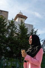 Portrait of cheerful muslim woman wearing a hijab isolated with mosque background. Vertical view of arabic woman drinking coffee outdoors. Muslim women lifestyle.