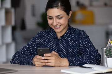 Wall Mural - Close up smiling Indian woman using smartphone at workplace, happy young businesswoman entrepreneur or student looking at phone screen, typing chatting online in social network, surfing internet